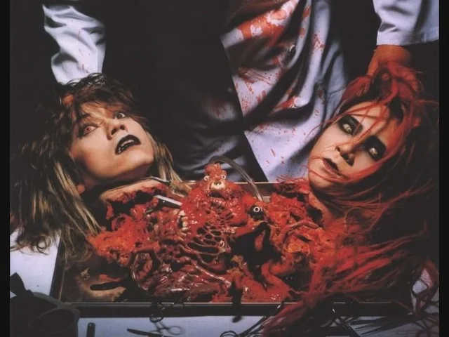 A closer view of the two severed-head prosthetics on the tray, with a figure in a bloodied lab coat standing behind them.