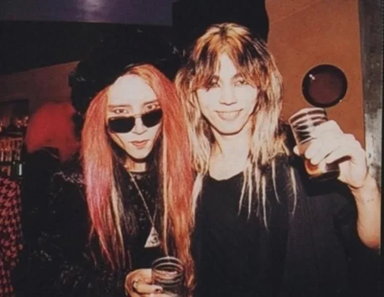 hide, in sunglasses and red hair, stands with Screaming Mad George at a bar, both holding drinks.