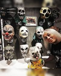 Screaming Mad George, grey-haired, leans in at the side of a lit display case holding around ten Slipknot masks.