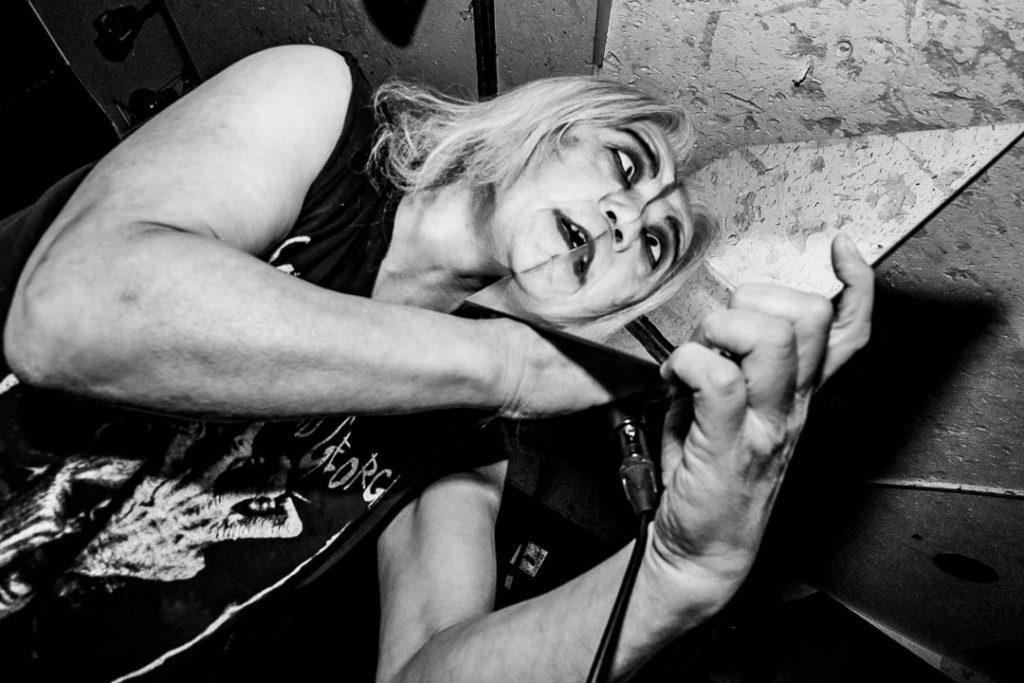 Black-and-white shot of Screaming Mad George mid-performance, white hair loose, gripping a microphone cable and shouting, shot from a low angle in a small venue.