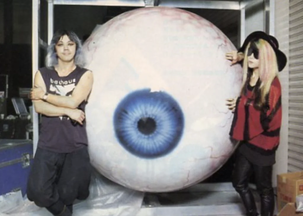 Screaming Mad George and hide stand on either side of a giant eyeball stage prop in a loading bay.