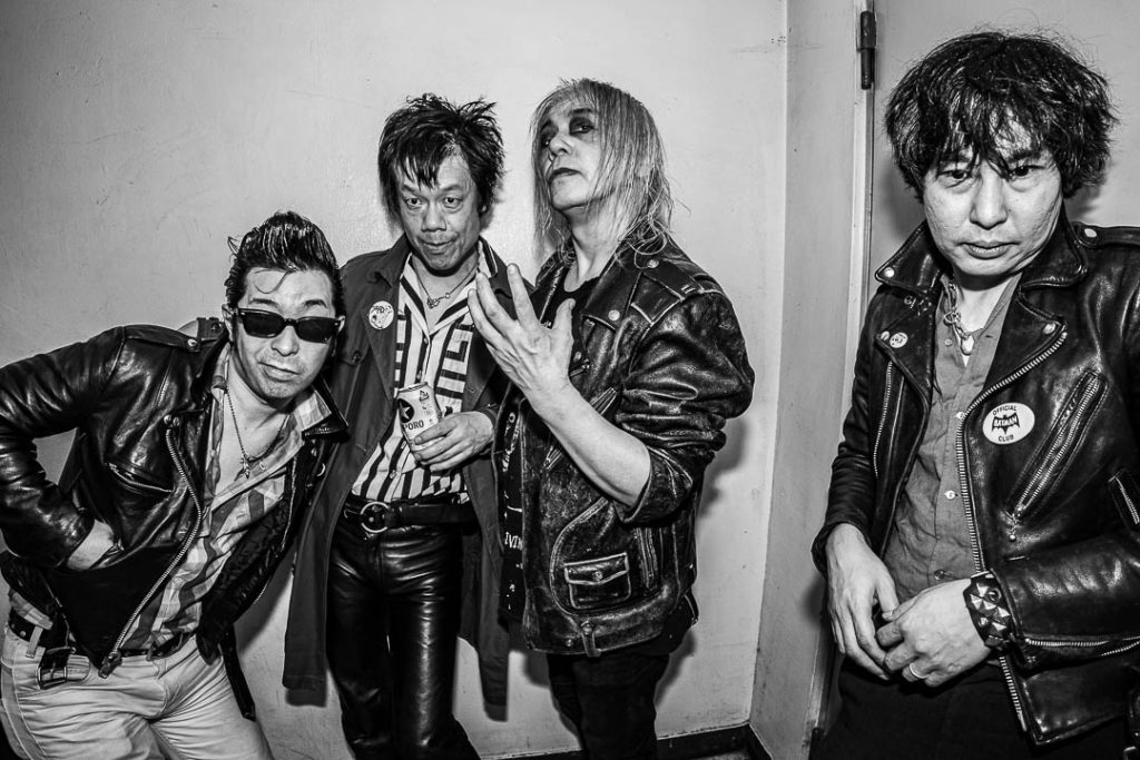 Black-and-white group photo of Screaming Mad George and three bandmates standing against a white backstage wall in leather jackets.
