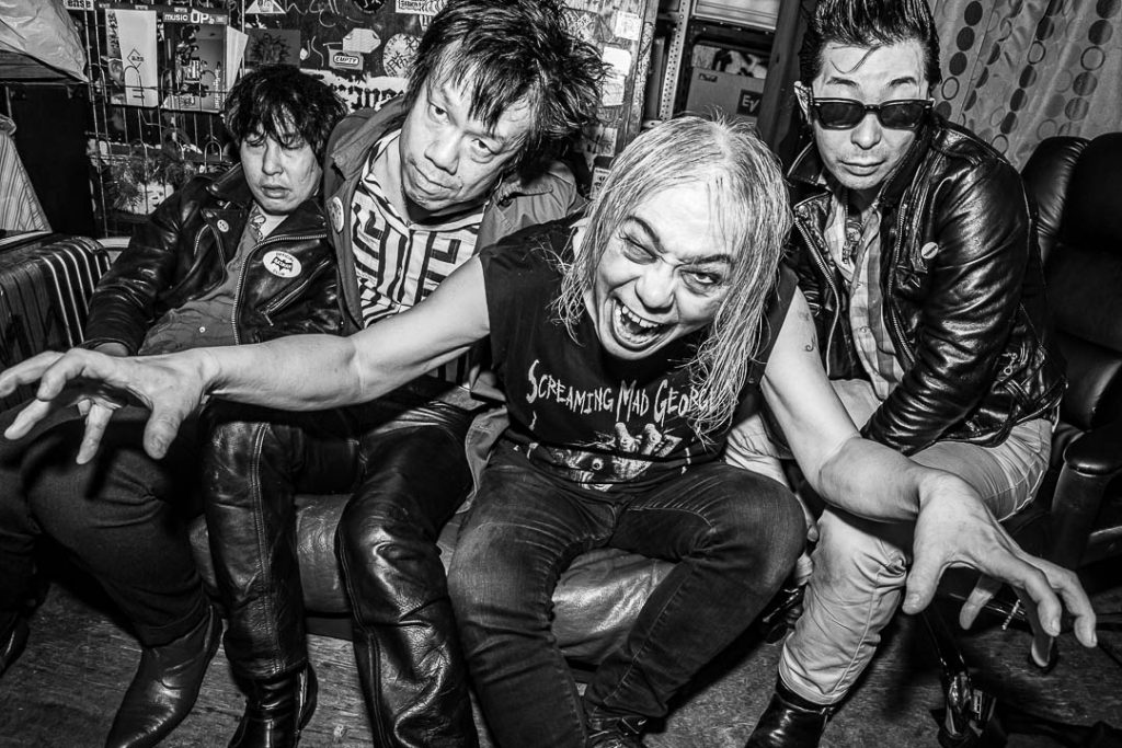 Black-and-white group photo: Screaming Mad George, in a shirt printed with his own name, grins open-mouthed on a couch with three bandmates in leather jackets.