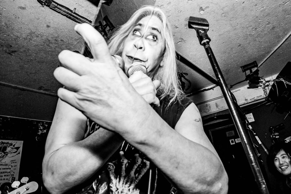 Black-and-white shot of Screaming Mad George singing into a handheld microphone with one arm thrust toward the camera in a low-ceilinged club.