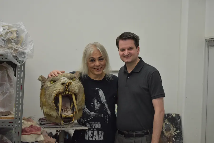 Screaming Mad George and Brett Homenick smile beside a saber-toothed creature head in George's Osaka workshop.