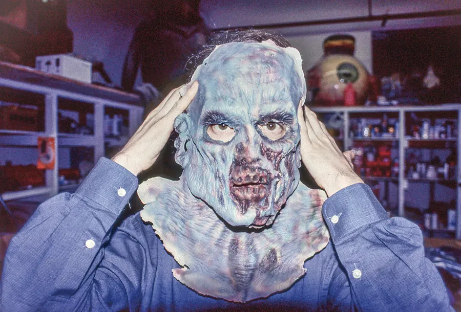 A person holds a zombie face-and-neck mask in front of studio shelves.