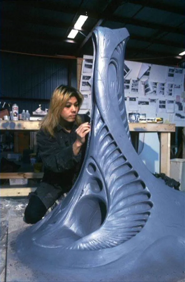 A long-haired artist works beside a tall ribbed creature-effects form in a workshop.