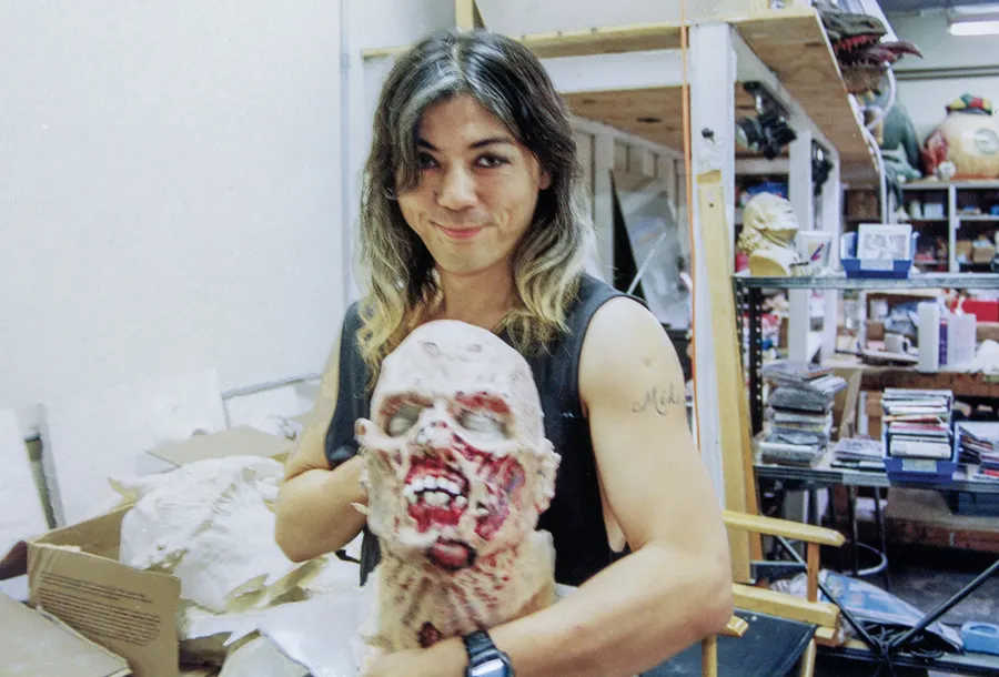 Screaming Mad George smiles while holding a severed-head prop in a studio crowded with creature sculptures.