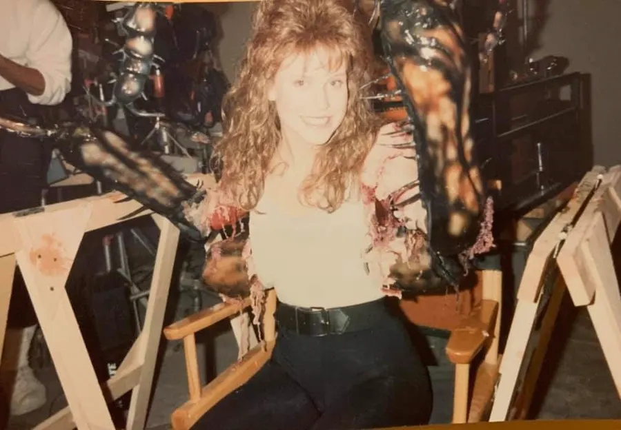 Brooke Theiss sits in a director's chair wearing the completed cockroach-arm appliances as crew stand nearby.