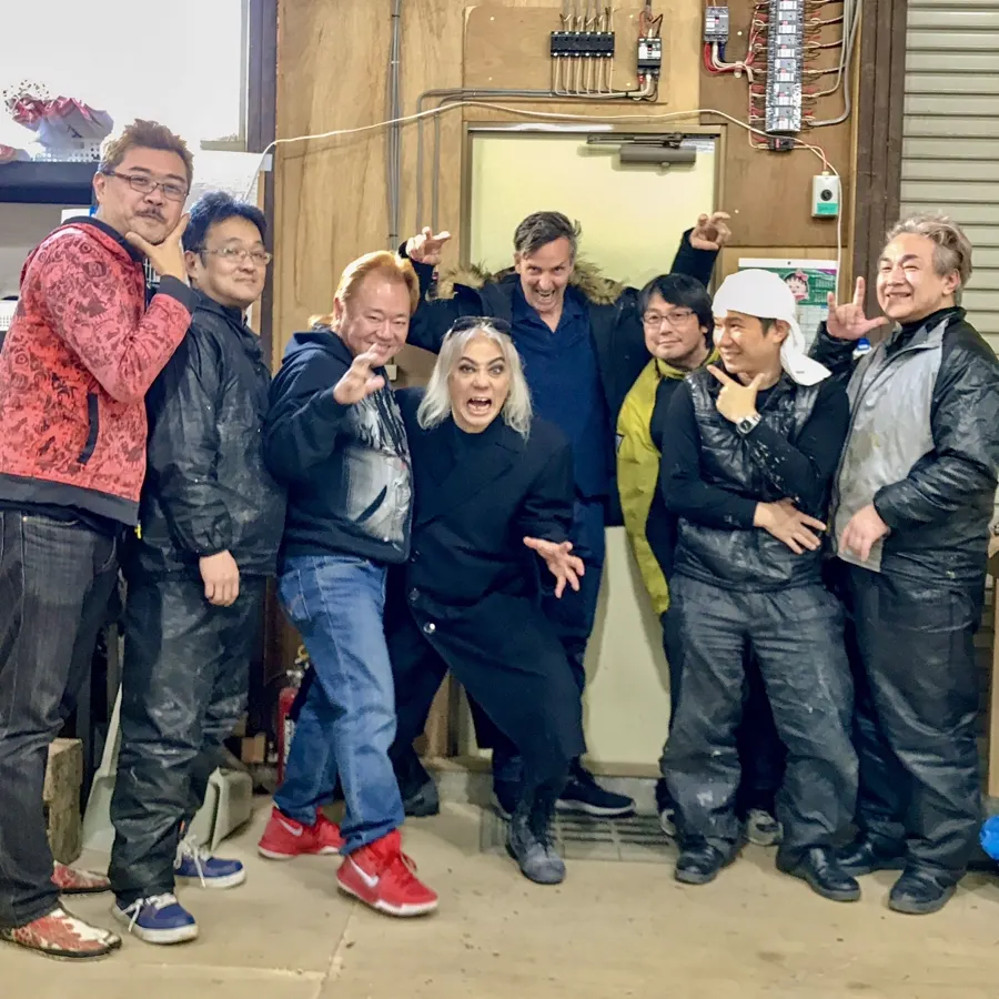 Screaming Mad George stands at the center of a playful group portrait in a workshop.